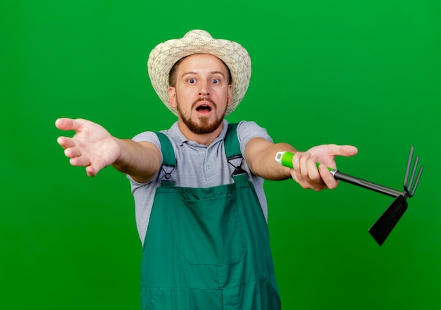 Free Photo impressed young handsome slavic gardener in uniform and hat looking  stretching out hands  holding rake isolated on green wall