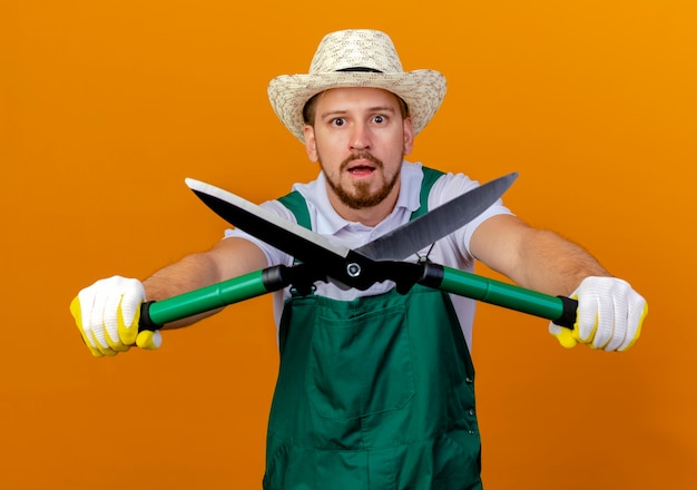 Free photo impressed young handsome slavic gardener in uniform wearing hat and gardening gloves stretching out pruners towards isolated