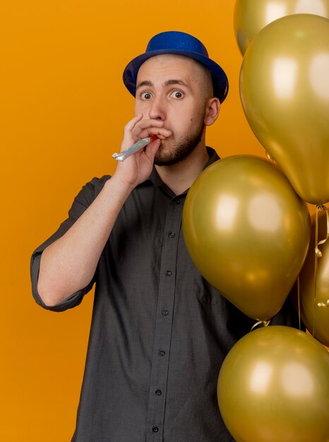 Impressed young handsome slavic party guy wearing party hat holding balloons blowing party blower looking at camera isolated on orange background