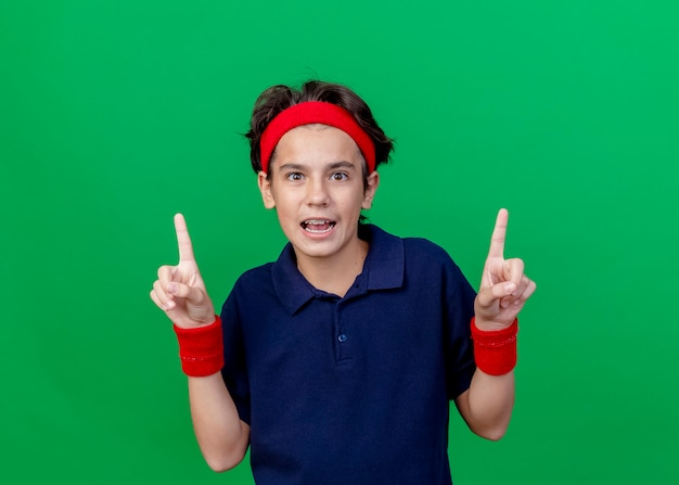 Impressed young handsome sporty boy wearing headband and wristbands with dental braces looking at camera pointing up isolated on green background