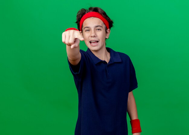 Impressed young handsome sporty boy wearing headband and wristbands with dental braces looking and pointing at camera isolated on green background with copy space