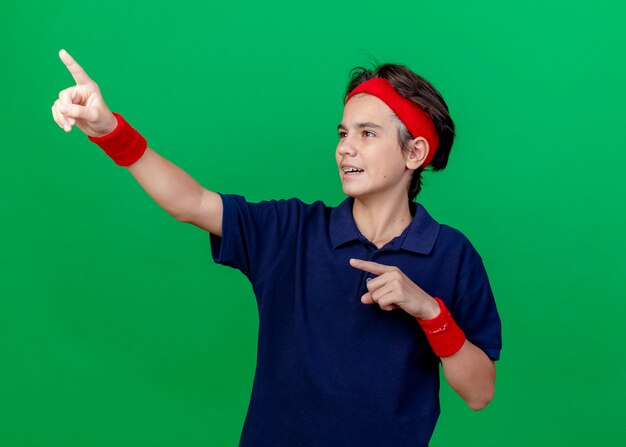 Impressed young handsome sporty boy wearing headband and wristbands with dental braces looking and pointing at side isolated on green background with copy space