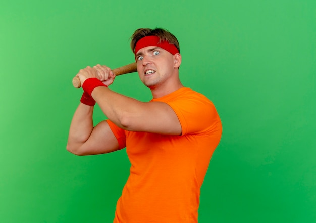 Impressed young handsome sporty man wearing headband and wristbands holding baseball bat getting ready to beat isolated on green