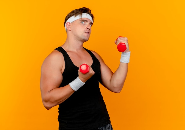 Impressed young handsome sporty man wearing headband and wristbands holding dumbbells isolated on orange