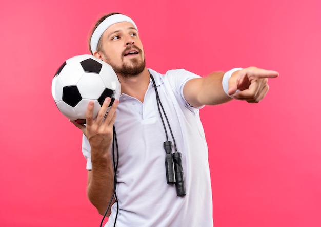 Free photo impressed young handsome sporty man wearing headband and wristbands holding soccer ball looking at side and pointing straight with jump rope around neck isolated on pink wall