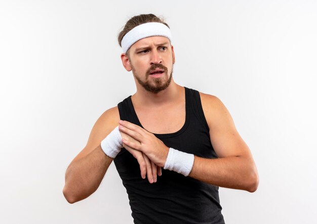 Impressed young handsome sporty man wearing headband and wristbands looking at side putting hand on another one isolated on white wall