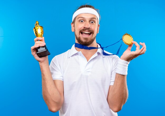 Impressed young handsome sporty man wearing headband and wristbands and medal around neck holding medal and winner cup  isolated on blue wall