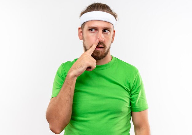 Impressed young handsome sporty man wearing headband and wristbands putting finger on nose looking at side isolated on white wall