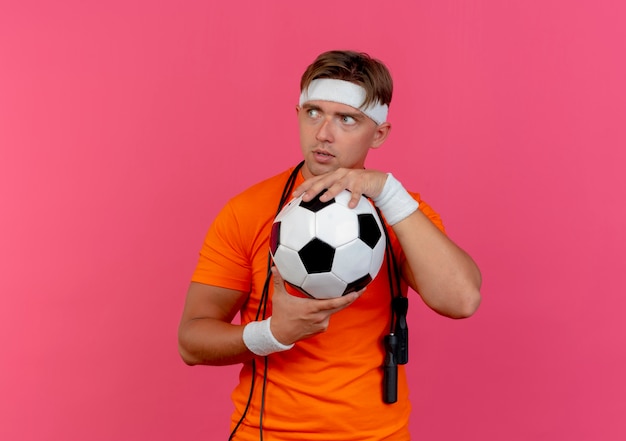 Free Photo impressed young handsome sporty man wearing headband and wristbands with jump rope around neck holding soccer ball looking at side isolated on pink