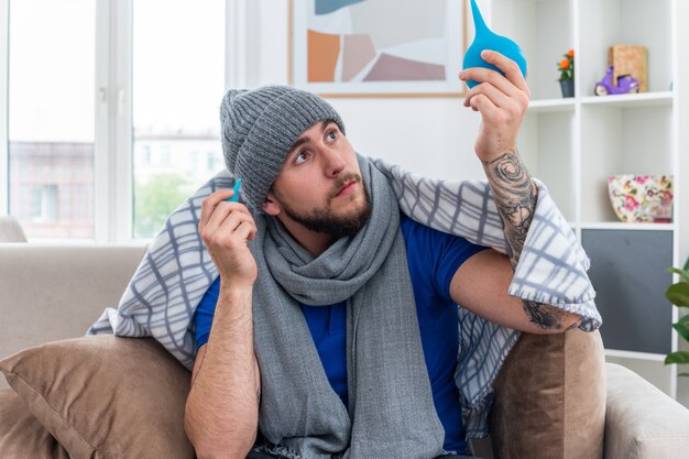 impressed young ill man wearing scarf and winter hat sitting on sofa in living room wrapped in blanket holding enemas raising one looking at it