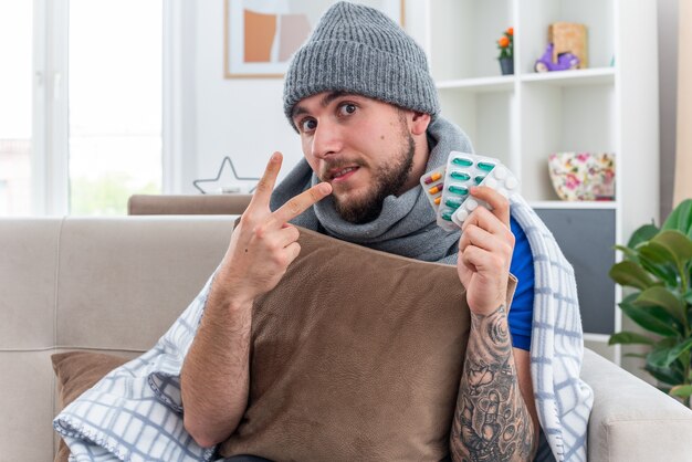 Impressed young ill man wearing scarf and winter hat wrapped in blanket sitting on sofa in living room holding pillow looking at camera showing packs of pills and showing two with hand