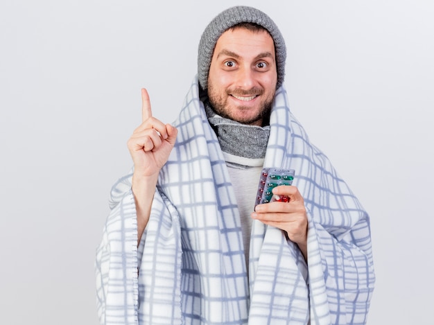 Impressed young ill man wearing winter hat and scarf wrapped in plaid holding pills