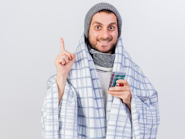 Impressed young ill man wearing winter hat and scarf wrapped in plaid holding pills