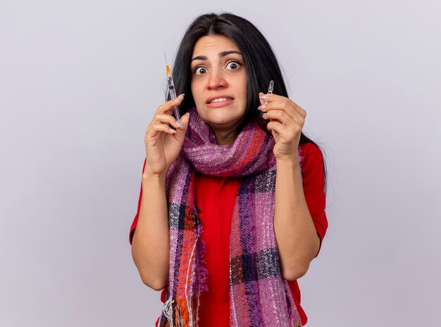 Impressed young ill woman wearing scarf holding syringe and ampoule looking at front isolated on white wall