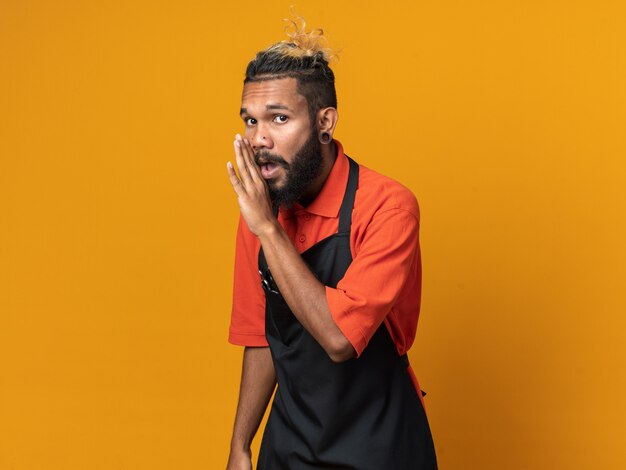 Impressed young male barber wearing uniform standing in profile view looking at front whispering isolated on orange wall with copy space