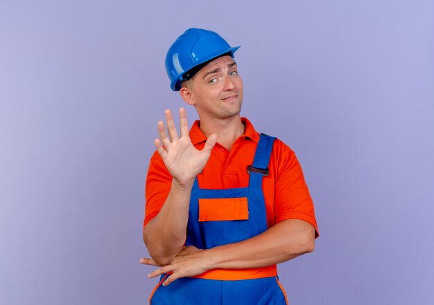 impressed young male builder wearing uniform and safety helmet showing five