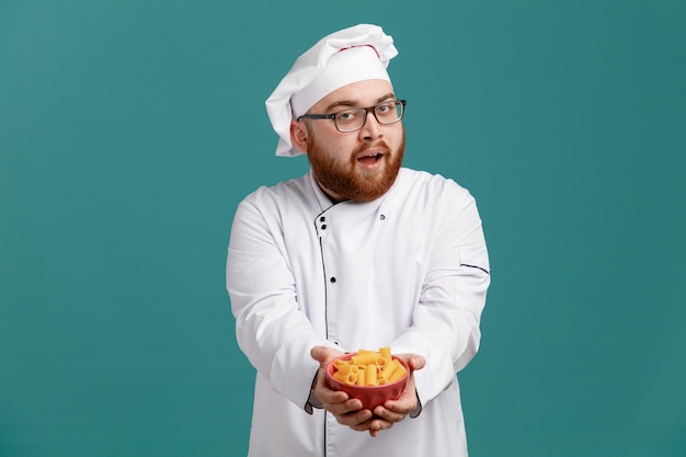 Impressed young male chef wearing glasses uniform and cap looking at camera stretching bowl of macaroni pasta out towards camera isolated on blue background