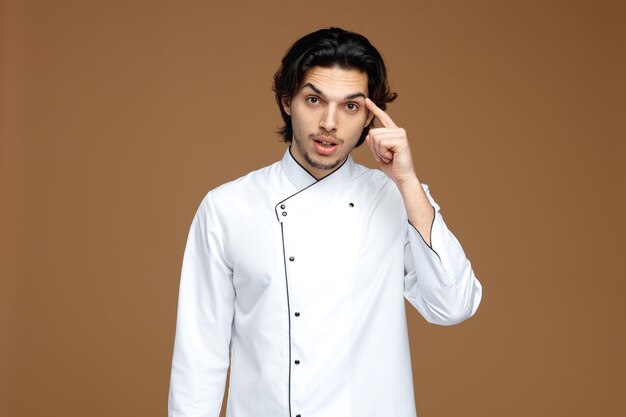 impressed young male chef wearing uniform looking at camera showing think gesture isolated on brown background