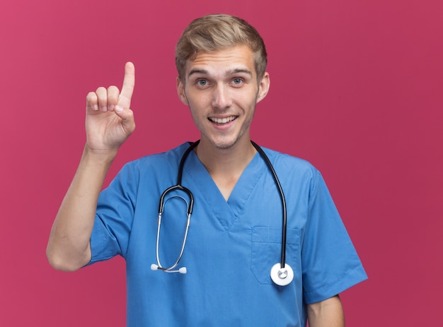 Impressed young male doctor wearing doctor uniform with stethoscope points at up isolated on pink wall