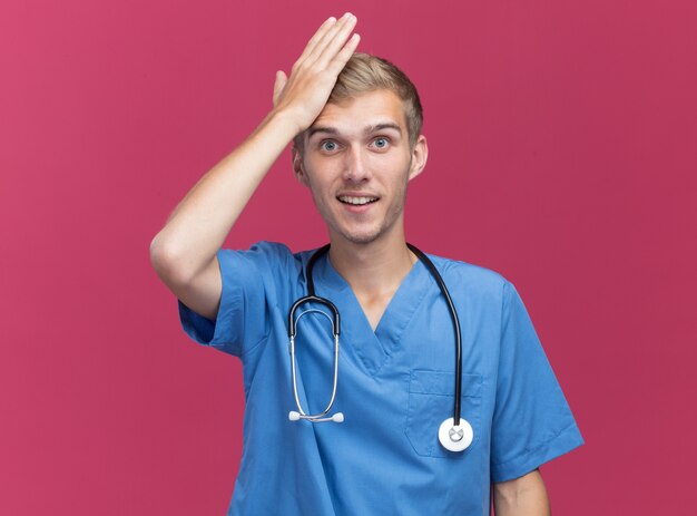 Free photo impressed young male doctor wearing doctor uniform with stethoscope putting hand on forehead isolated on pink wall