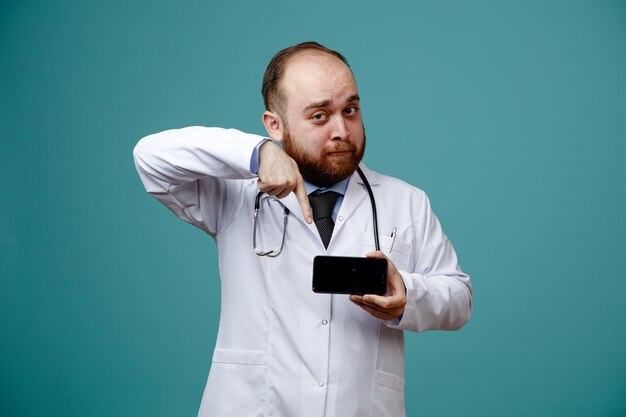 Impressed young male doctor wearing medical coat and stethoscope around his neck showing mobile phone pointing at it looking at camera isolated on blue background