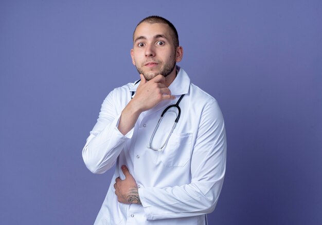 Impressed young male doctor wearing medical robe and stethoscope around his neck putting hand on belly and touching chin isolated on purple