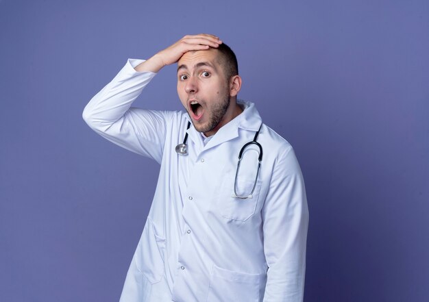 Impressed young male doctor wearing medical robe and stethoscope around his neck putting hand on head isolated on purple