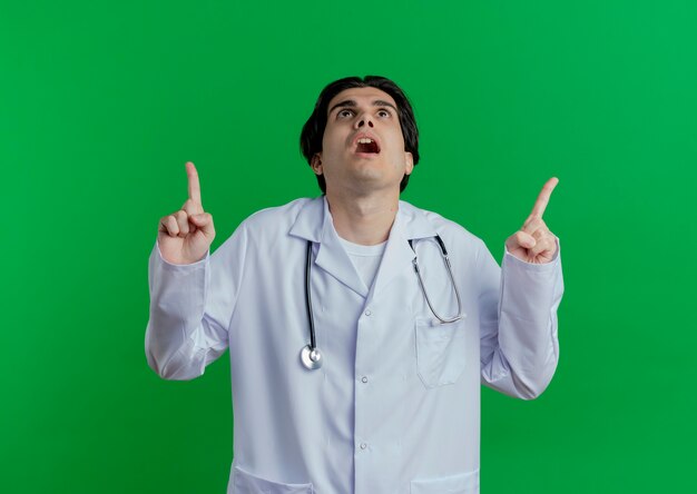 Impressed young male doctor wearing medical robe and stethoscope looking and pointing up isolated on green wall with copy space