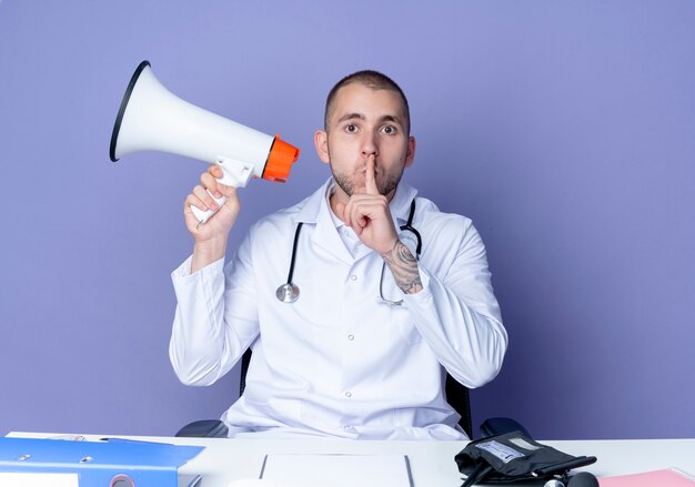 Impressed young male doctor wearing medical robe and stethoscope sitting at desk with work tools holding speaker and gesturing silence isolated on purple