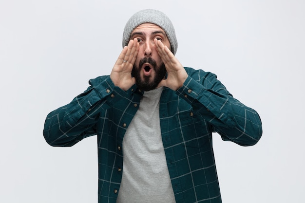 Impressed young man wearing winter hat looking at camera keeping hands near mouth calling someone out isolated on white background