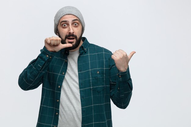 Impressed young man wearing winter hat looking at camera pointing to side isolated on white background