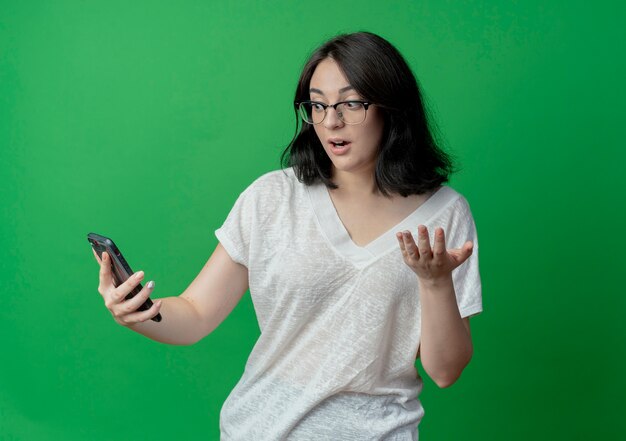 Impressed young pretty caucasian girl wearing glasses holding and looking at mobile phone and showing empty hand isolated on green background