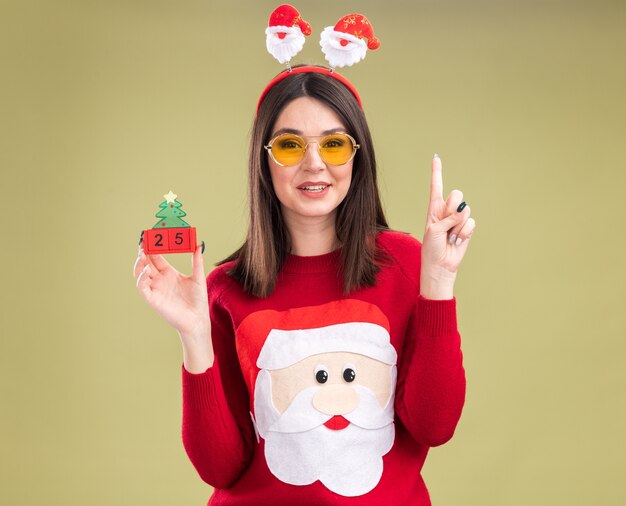 Impressed young pretty caucasian girl wearing santa claus sweater and headband with glasses holding christmas tree toy with date 