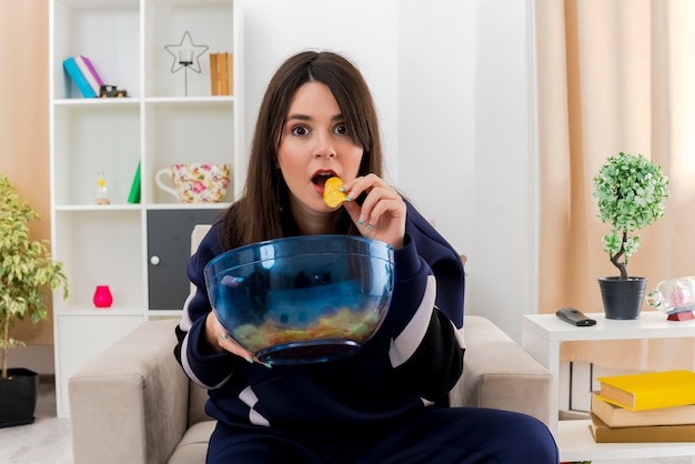 Impressed young pretty caucasian woman sitting on armchair in designed living room holding bowl of chips looking and eating potato chip