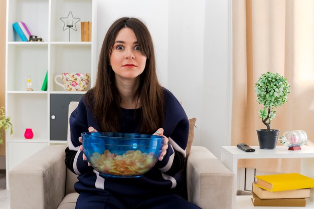 Impressed young pretty caucasian woman sitting on armchair in designed living room holding bowl of chips and looking