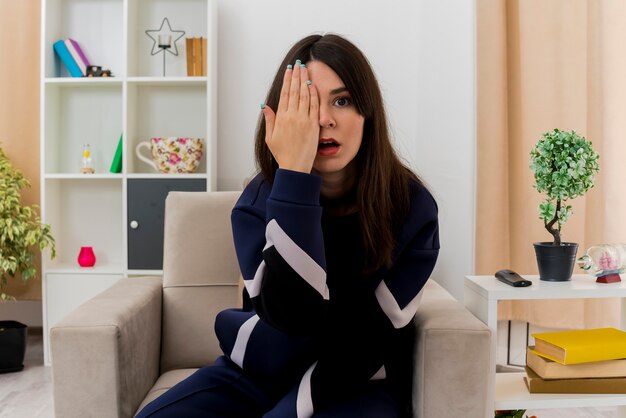 Impressed young pretty caucasian woman sitting on armchair in designed living room putting hand on face covering half of face looking