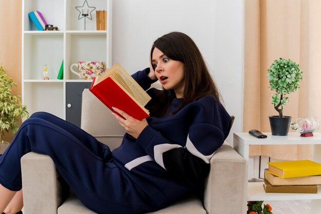 Impressed young pretty caucasian woman sitting on armchair in designed living room reading book putting hand on head