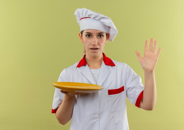 Impressed young pretty cook in chef uniform holding empty plate and raising hand isolated on green wall