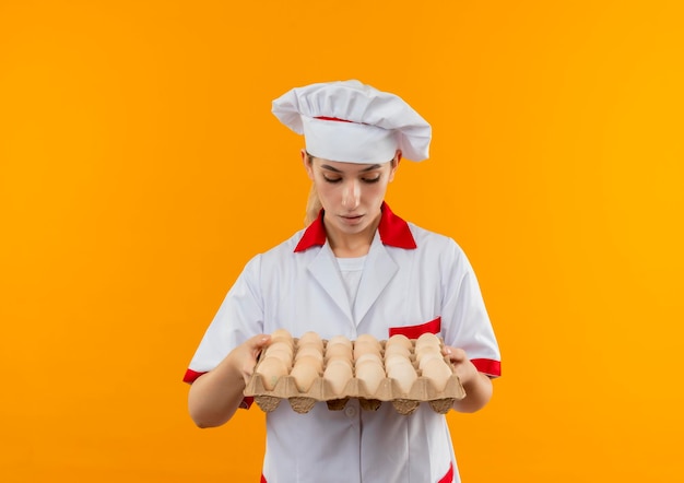 Impressed young pretty cook in chef uniform holding and looking at carton of eggs isolated on orange wall