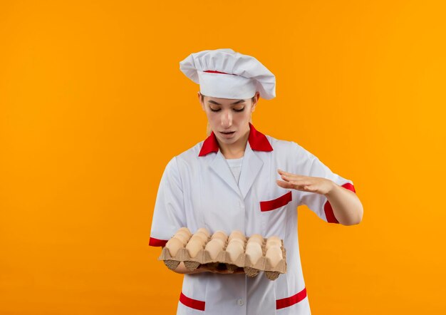 Impressed young pretty cook in chef uniform holding and looking at carton of eggs keeping hand on air isolated on orange wall with copy space