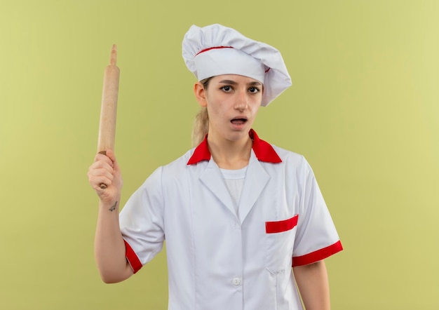 Impressed young pretty cook in chef uniform holding rolling pin isolated on green wall