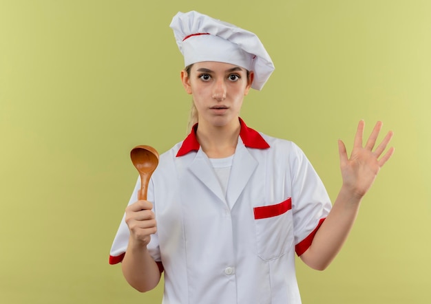 Impressed young pretty cook in chef uniform holding spoon showing empty hand isolated on green wall