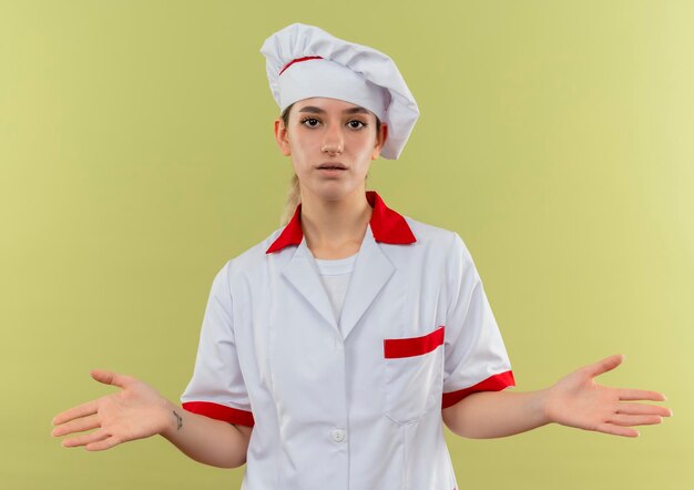 Impressed young pretty cook in chef uniform showing empty hands isolated on green wall