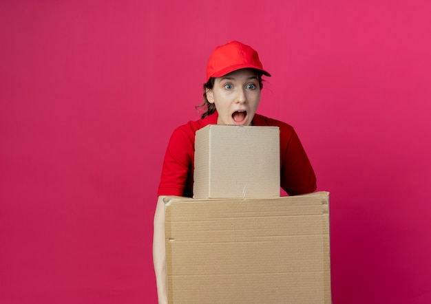 Free Photo impressed young pretty delivery girl in red uniform and cap holding carton boxes looking straight isolated on crimson background with copy space