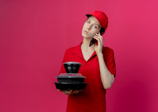 Free photo impressed young pretty delivery girl wearing red uniform and cap talking on phone and holding food containers looking at camera isolated on crimson background with copy space