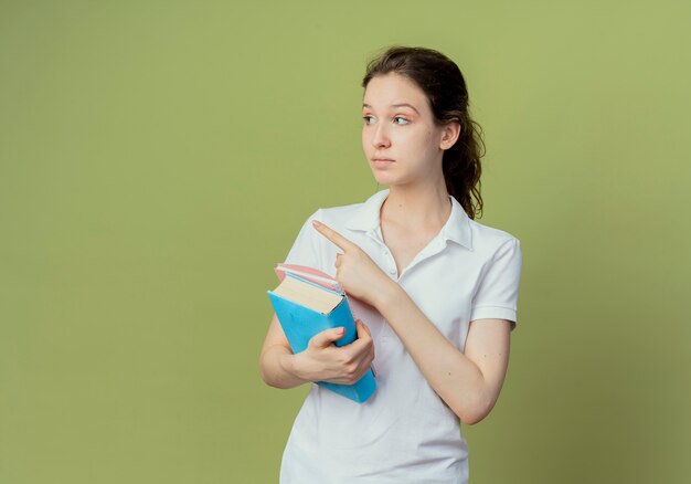 Impressed young pretty female student holding book and note pad looking and pointing at side isolated on olive green background with copy space