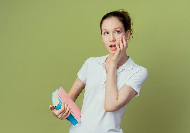 Impressed young pretty female student holding book and note pad putting hand on face and looking at side isolated on olive green background with copy space