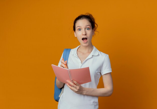 Impressed young pretty female student wearing back bag holding pen and note pad isolated on orange background with copy space