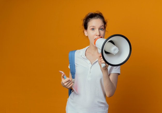 Impressed young pretty female student wearing back bag holding pen and note pad talking by speaker isolated on orange background with copy space