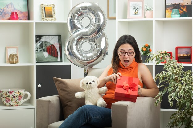 Impressed young pretty girl in optical glasses opening and looking at gift box sitting on armchair in living room on march international women's day